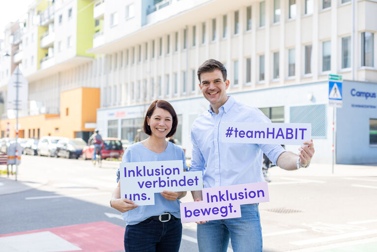 Andreas Kauba und Cornelia Girmindl (Geschäftsführungsteam von HABIT) stehen an einer Straßenecke und halten Schilder hoch (Inklusion verbindet und Inklusion bewegt).
