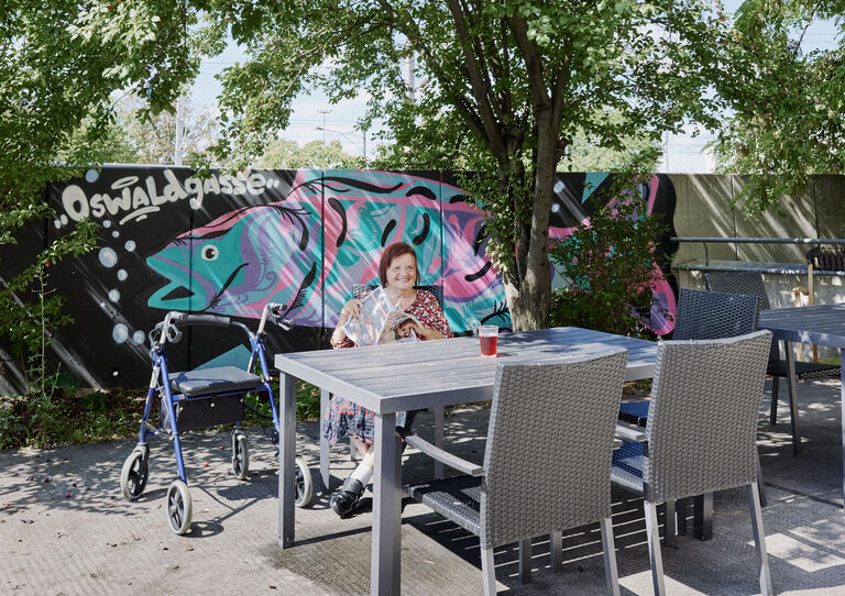 Kundin sitzt bei Tisch im Garten der HABIT WG Oswaldgasse, im Hintergrund ist ein Graffiti mit Fisch