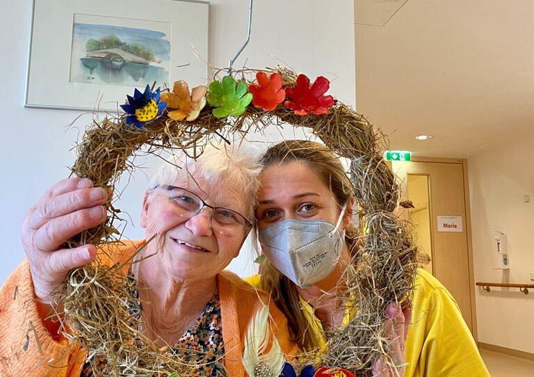 Bewohnerin und Mitarbeiterin blicken durch den selbst gebastelten Kranz.