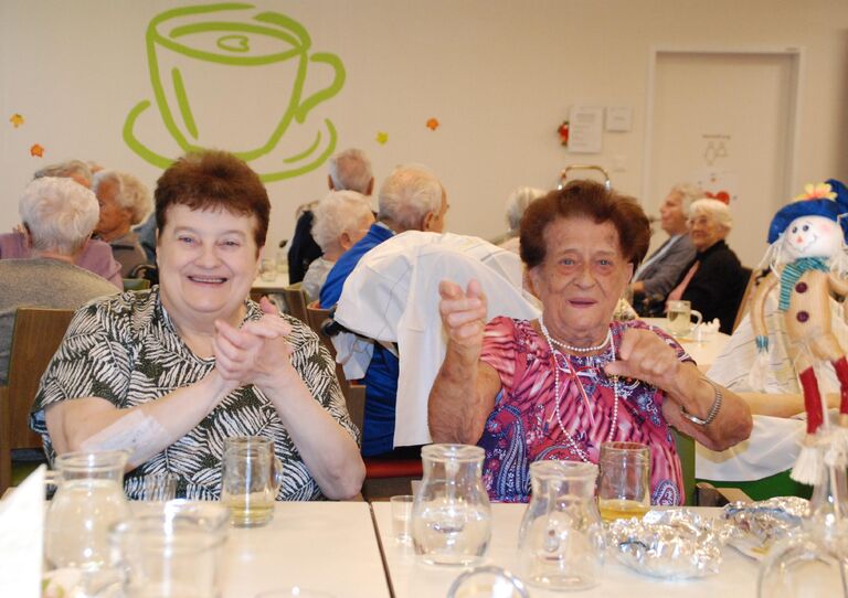 Zwei Bewohnerinnen des Stadtheims sitzen nebeneinander am Tisch, lachen fröhlich und klatschen.