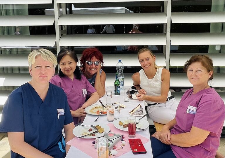 Kolleg*innen sitzen am Esstisch outdoor zusammen und lachen in die Kamera.