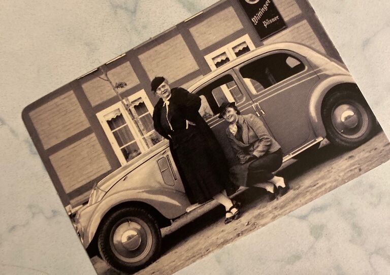 Schwarz-Weiß Foto von zwei Damen im Mantel, die vor einem Käfer posieren