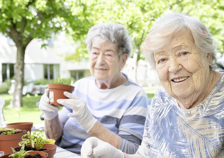 Zwei Bewohnerinnen bei der Gartenarbeit, eine lächelt in die Kamera Zwei Bewohnerinnen bei der Gartenarbeit, eine lächelt in die Kamera