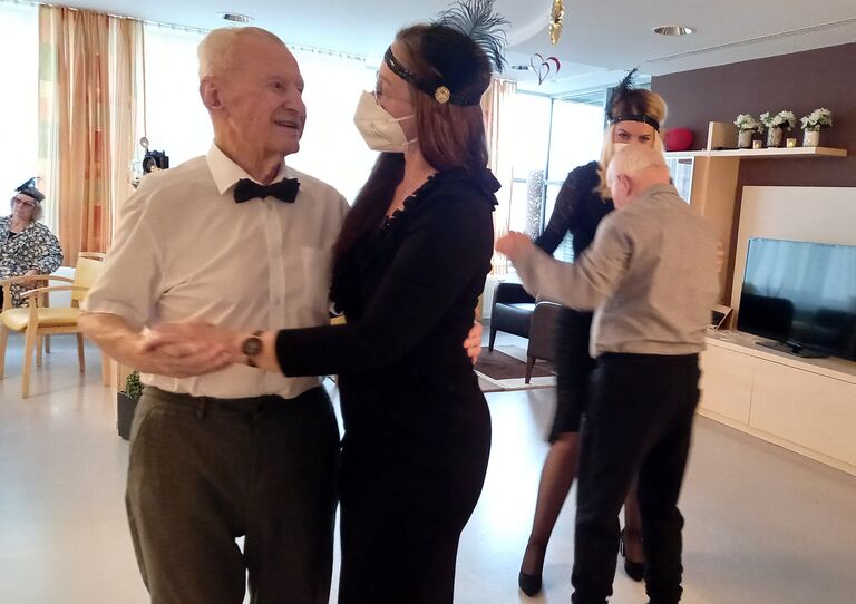 Eine Mitarbeiterin tanzt mit einem Bewohner. Beide sind elegant angezogen. Die Mitarbeiterin trägt einen Kopfschmuck aus den zwanziger Jahren.