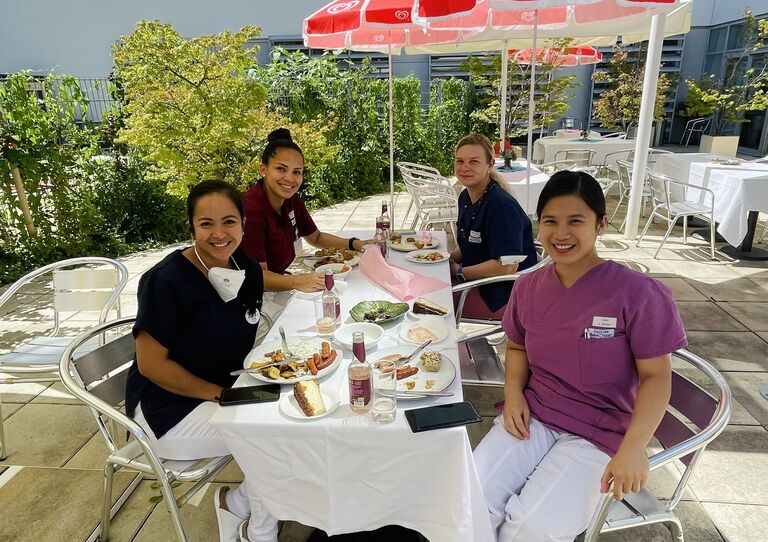 Kolleg*innen sitzen am Esstisch outdoor zusammen und lachen in die Kamera.