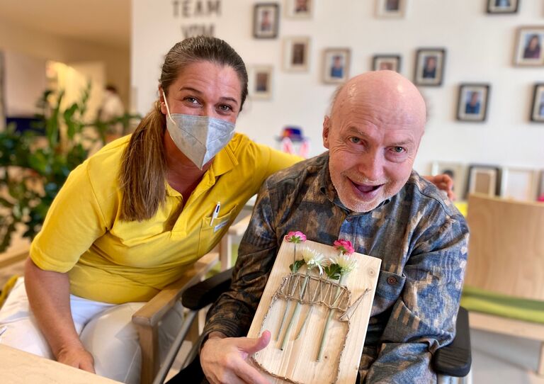 Mitarbeiterin sitzt neben Bewohner und er hält sein Bild hoch.