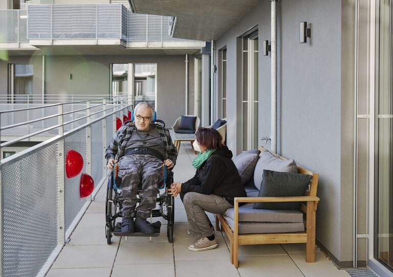 Ein Kunde im Rollstuhl und eine Mitarbeiterin sitzend daneben befinden sich beide auf der Terrasse des GV Verbundes.