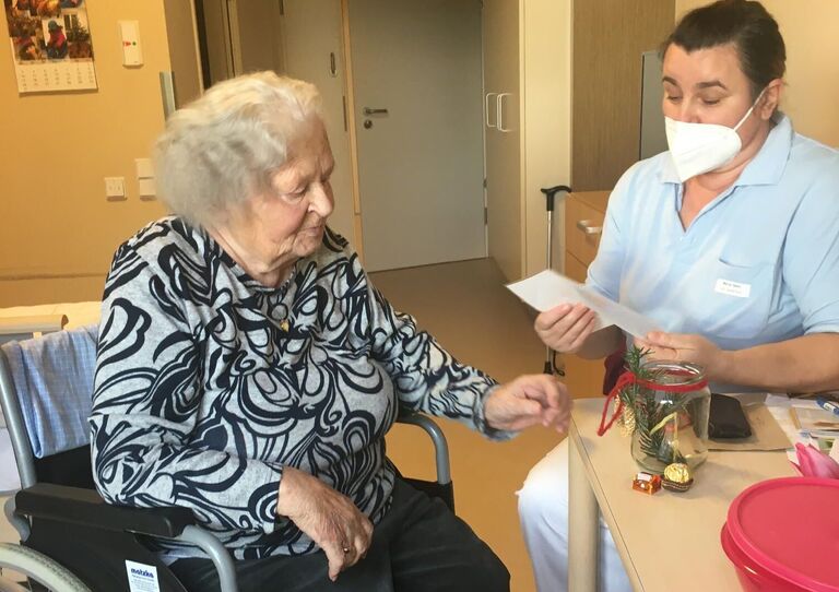Seniorenbetreuerin Marija liest einer Bewohnerin, die am Tisch sitzt etwas vor.