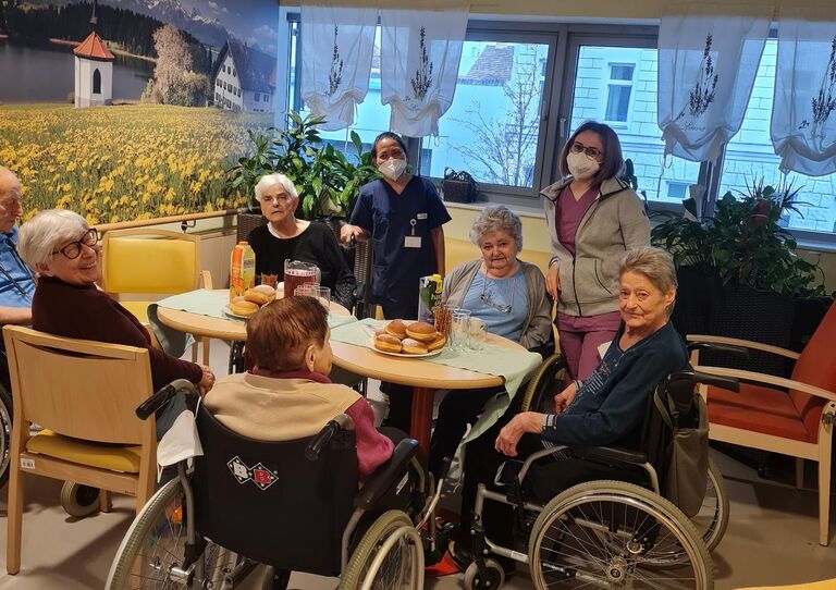 Bewohner*innen und Mitarbeiter*innen stehen und sitzen rund um den Tisch und blicken in die Kamera.