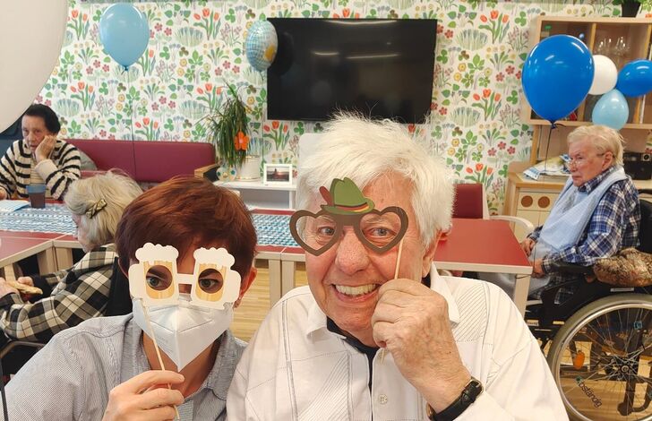 Eine Mitarbeiterin und ein Bewohner nebeneinander bei einem kleinen Oktoberfest. Sie tragen lustige Papiermasken, die zum Motto passen.