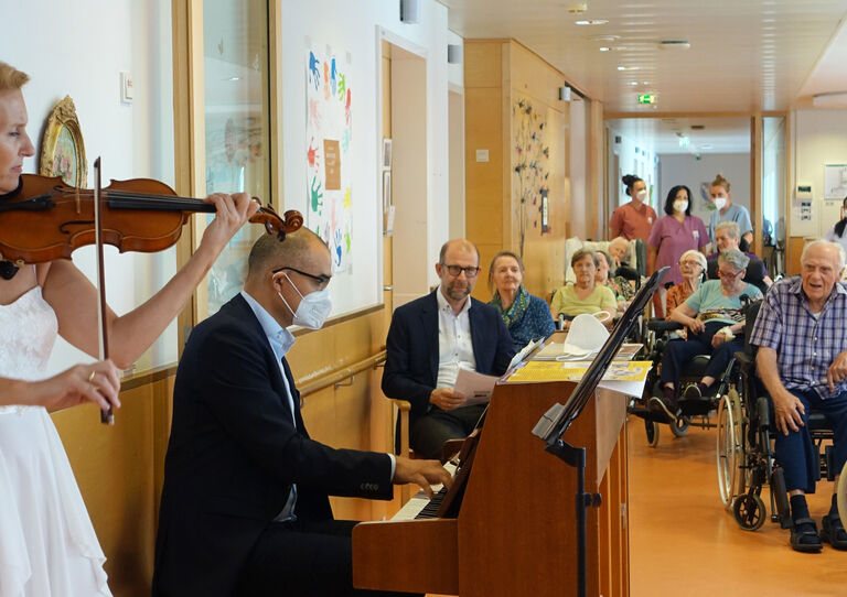Ärztlicher Leiter Tokiostraße und seine Ehefrau am Klavier und an der Geige. Die Bewohner*innen sehen interessiert zu.