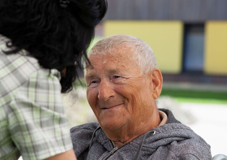 Ein Bewohner im Rollstuhl blickt eine Mitarbeiterin, die von hinten zu sehen ist, mit großer Zuneigung an und hält sie bei den Armen.