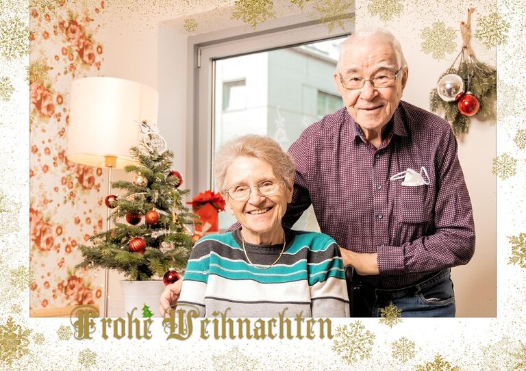 Eine Bewohnerin wird im Sitzen von einem Bewohner lieb von hinten seitlich umarmt, sie blicken beide freundlich in die Kamera und befinden sich vor einer weihnachtlichen Kulisse.