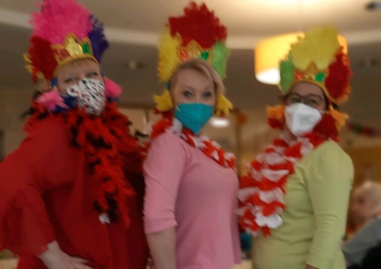 Drei Mitarbeiter*innen haben bunte Masken und einen bunten Kopfschmuck und lachen in die Kamera.