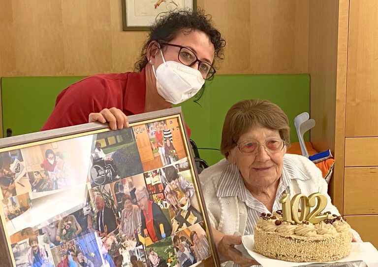 Mitarbeiterin hält ein Bild mit vielen Fotos der Bewohnerin, Bewohnerin hält eine Geburtstagstorte in der Hand zum 102. Geburtstag.
