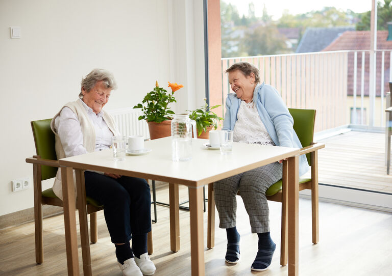 Zwei Bewohnerinnen sitzen an einem Tisch und lachen herzlich miteinander.
