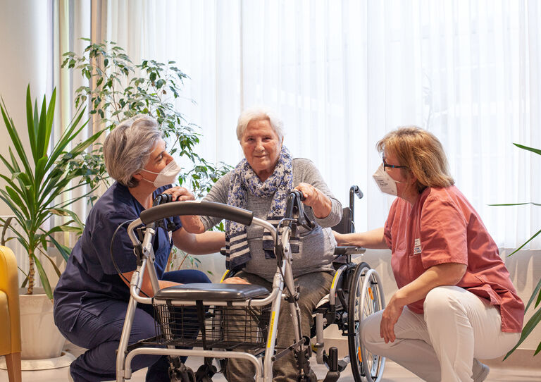 Zwei Mitarbeiterinnen knien neben einer Bewohnerin in der Mitte, die in die Kamera lächelt und sich auf die Gehhilfe stützt.