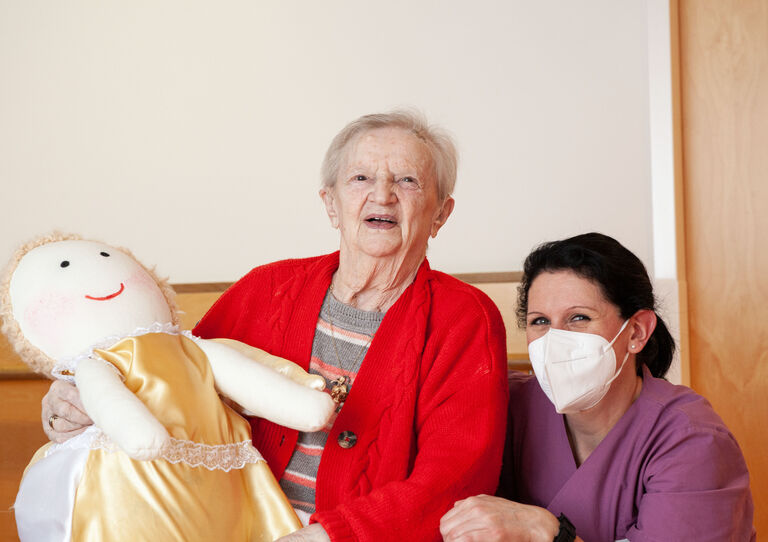 Bewohnerin mit ihrer Stoffpuppe und Mitarbeiterin kniend daneben.