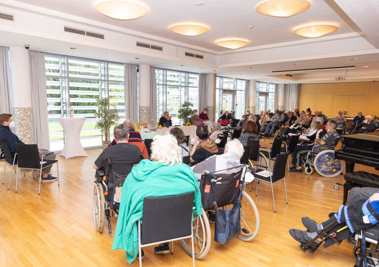 Aufnahme aus dem Festsaal, Bewohner*innen von hinten, ganz vorne sitzt Adi Hirschal