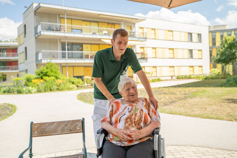 Raphael Herbst steht hinter einer Bewohnerin, die freundlich in die Kamera lächelt und schiebt ihren Rollstuhl.
