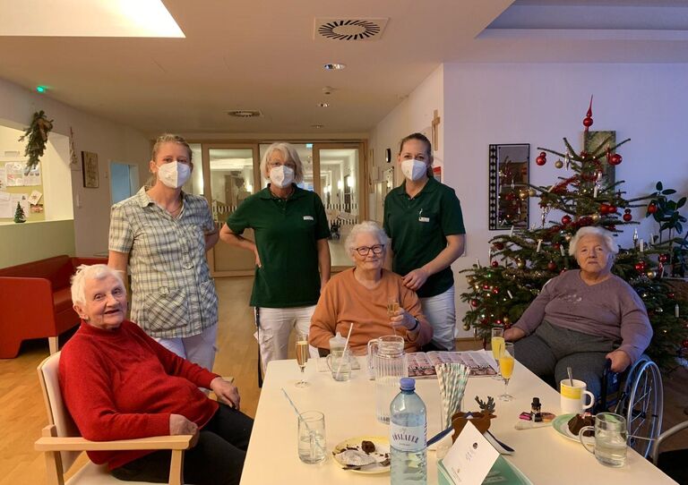 Einige Bewohnerinnen und einige Mitarbeiterinnen stehen rund um den Tisch mit einem Sektglas in der Hand.