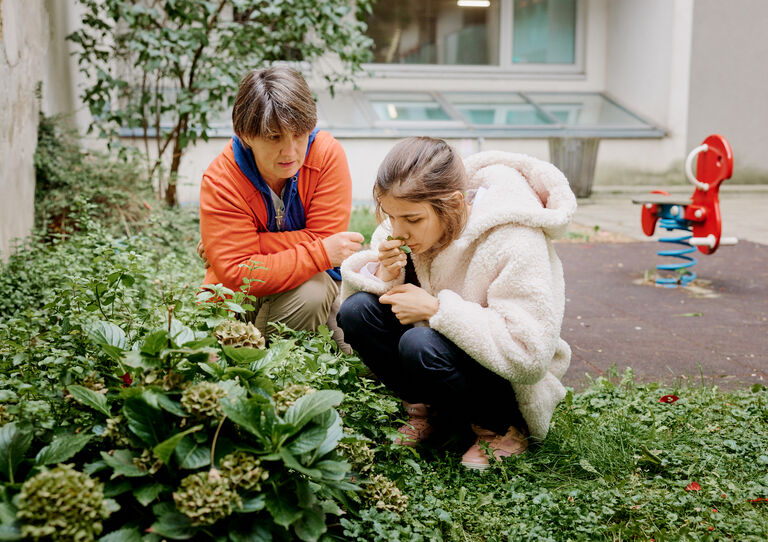 BTZ Lobenhauerngasse: Innenhof: eine Kundin knieht bei den Blumen und riecht daran, eine Mitarbeiterin sitzt neben ihr. 
