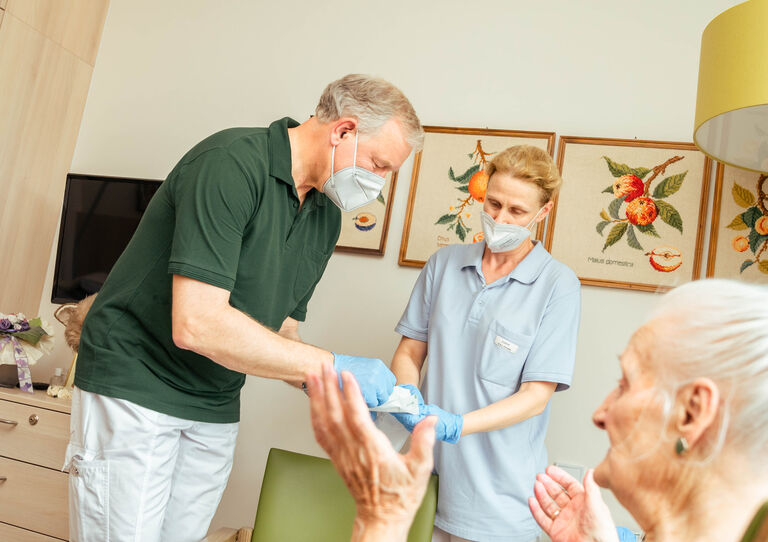 Landesrat Martin Eichtinger legt einer Bewohnerin einen Verband an, neben ihm steht eine HB Mitarbeiterin.