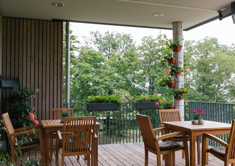 Die Terrasse einer Hausgemeinschaft mit Geranien, Holzstühlen und Holztischen.