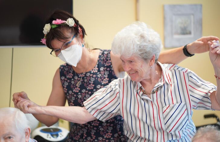 Eine Bewohnerin wird von einer Mitarbeiterin mit einem Haarkranz von hinten an den Händen gehalten und sie tanzen.
