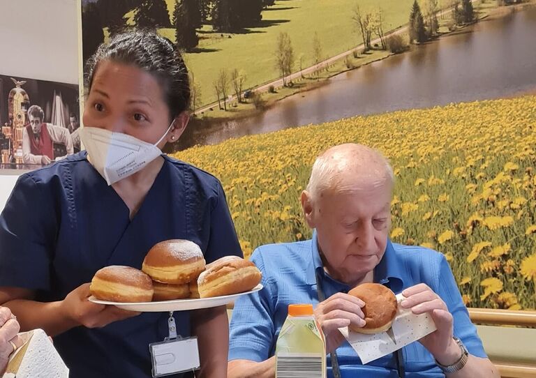 Eine Mitarbeiterin hält einen Teller mit Krapfen, von dem ein Bewohner einen genommen hat und ihn begutachtet.