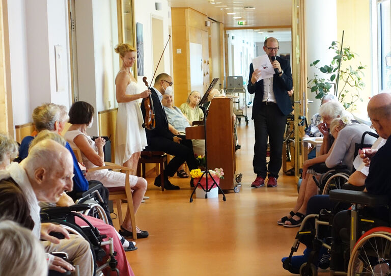Ärztlicher Leiter Tokiostraße und seine Ehefrau am Klavier und an der Geige. Die Bewohner*innen sehen interessiert zu.