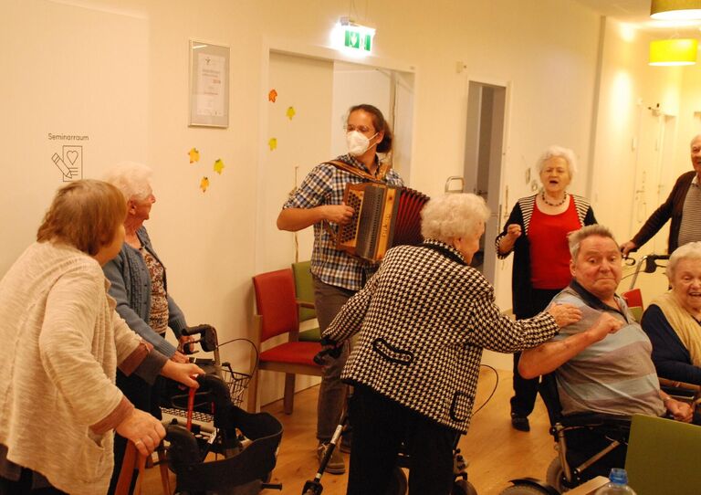 Die Bewohner*innen scharren sich rund um den Musikanten, der Ziehharmonika spielt und singen mit.