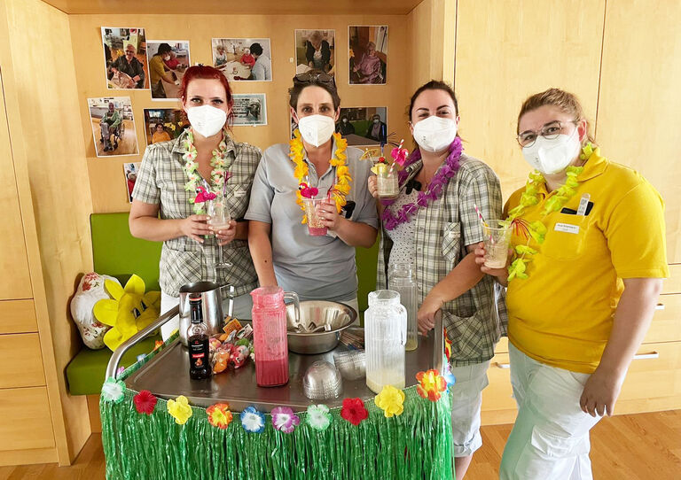 Vier Mitarbeiter*innen stehen hinter der mobilen Tiki Bar und halten einen Cocktail in den Händen.