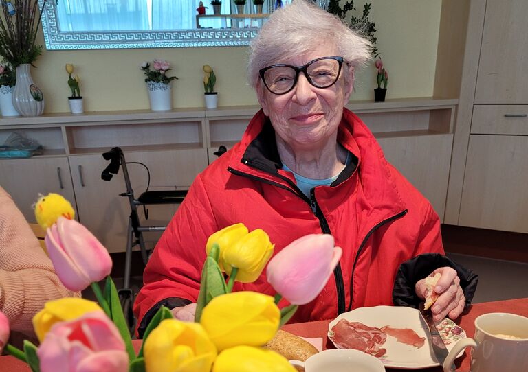 Eine Bewohnerin sitzt beim Frühstückstisch und lächelt in die Kamera.