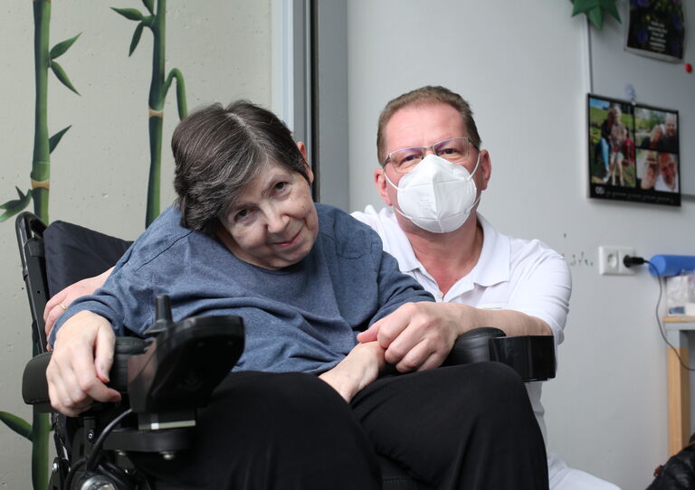 Eine Bewohnerin im Rollstuhl neigt ihren Kopf bedingt durch eine MS-Erkrankung stark nach links und lächelt in die Kamera. Ein Mitarbeiter sitzt hinter ihr und umarmt sie von hinten.