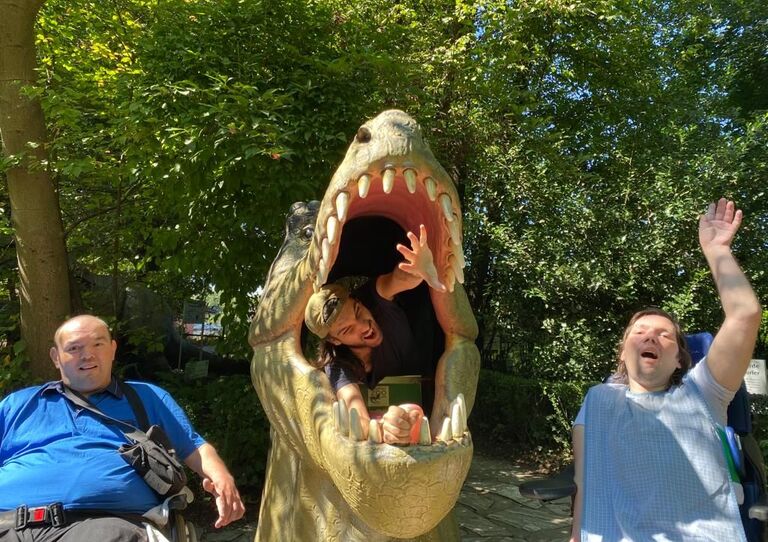 Zwei Kund*innen von HABIT sitzen neben einer Krokodilfigur und strecken die Hände in die Höhe. Ein Kunde streckt seinen Kopf von hinten durch das Krokodil durch und lacht.