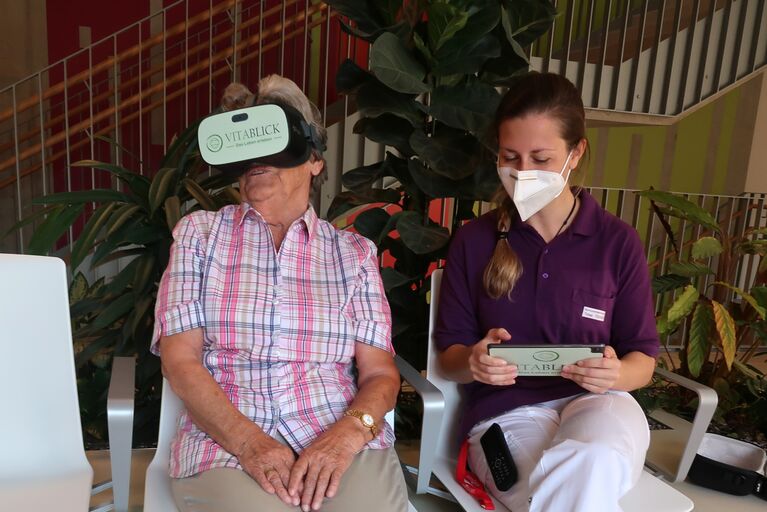 Bewohnerin mit VR-Brille neben Mitarbeiterin mit Beschreibung des besuchten Ortes.