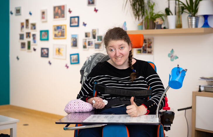 Haus der Barmherzigkeit/Ludwig Schedl Kundin im Aufenthaltsraum im Rollstuhl sitzend lächelt in die Kamera und streckt den Daumen nach oben.