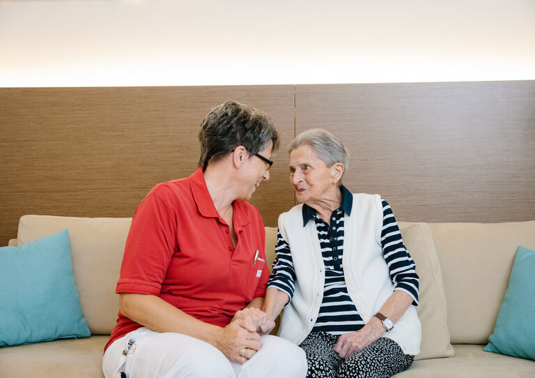 Mitarbeiterin und Bewohnerin sitzen nebeneinander auf der Couch, halten sich an den Händen und lächeln sich an Mitarbeiterin und Bewohnerin sitzen nebeneinander auf der Couch, halten sich an den Händen und lächeln sich an