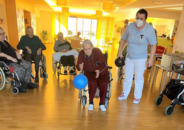 Eine Bewohnerin im Rollstuhl wirft den Ball. Betreuerin Marija verfolgt das Geschehen.