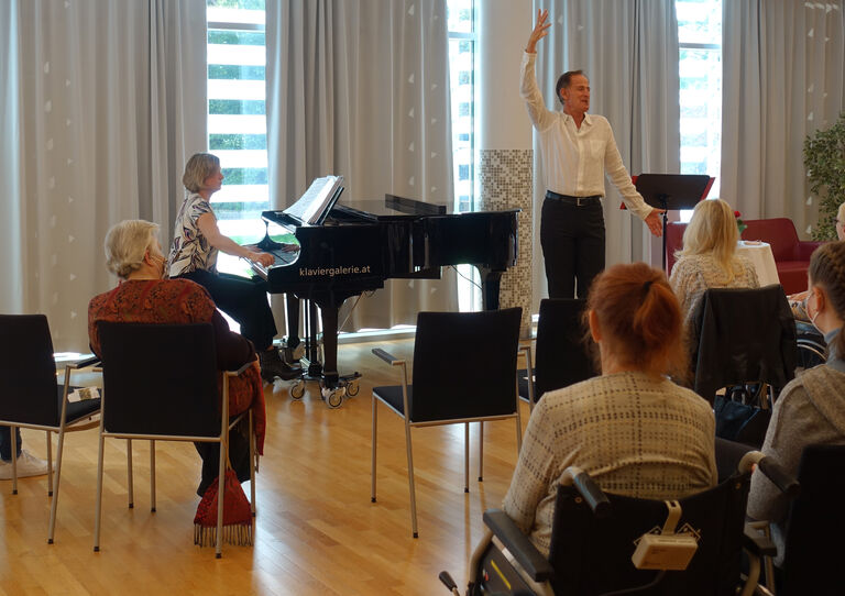 René Rumpold singt und streckt die Hand in die Höhe, links daneben steht ein Klavier und davor sitzt das Publikum.