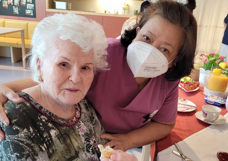 Bewohnerin und Mitarbeiterin lächeln freundlich in die Kamera, die Mitarbeiterin legt ihren Arm um die Bewohnerin.