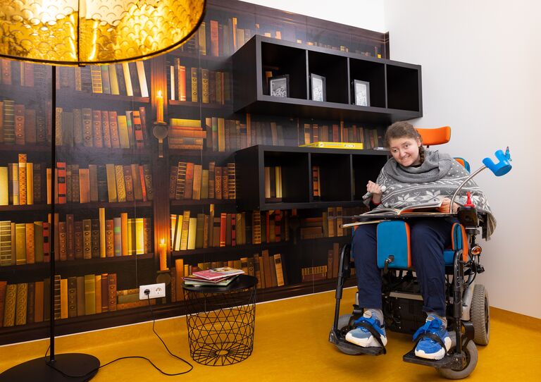 Eine Kundin im Rollstuhl sitzt vor einer Büchertapete und lächelt fröhlich in die Kamera.