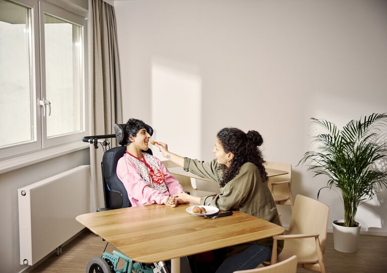Mitarbeiterin sitzt mit Kundin am Tisch und füttert sie. Mitarbeiterin sitzt mit Kundin am Tisch und füttert sie.