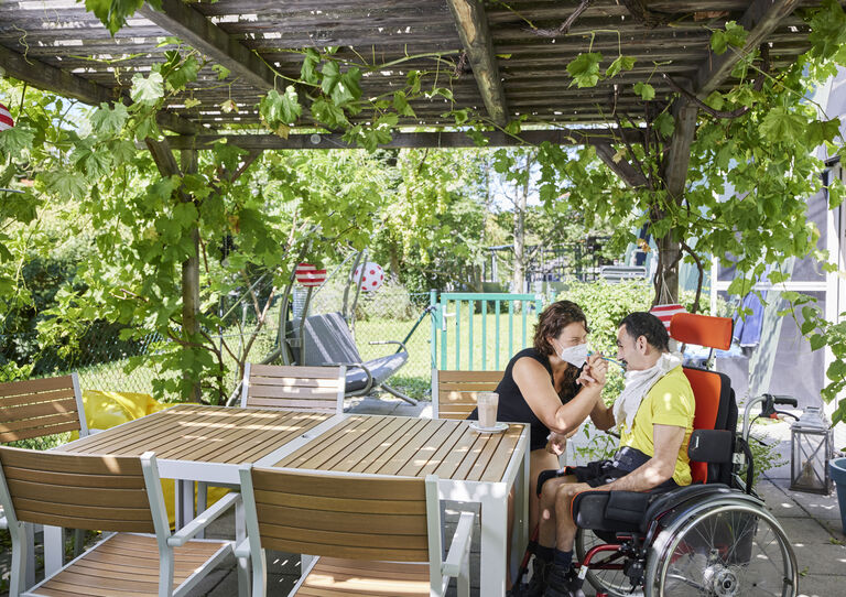 Kunde im Rollstuhl bekommt von Mitarbeiterin etwas zu Trinken auf der Terrasse mit Weinlaube in der HABIT WG Otto-Herschmann-Gasse