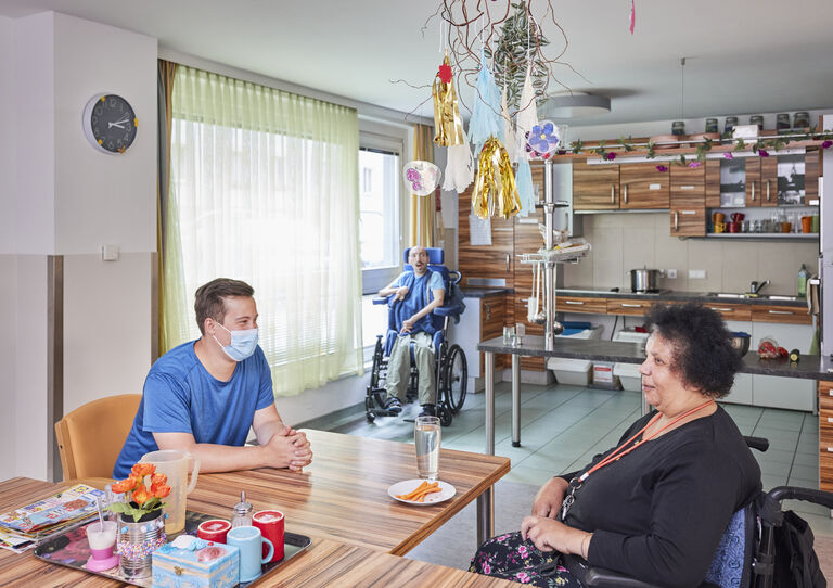 WG Rotenhofgasse eine Kundin und ein Mitarbeiter unterhalten sich am Tisch in der Küche, ein Kunde im Rollstuhl sieht aus dem Fenster