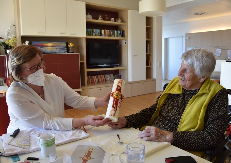 Eine Mitarbeiterin reicht einer Bewohnerin eine selbst gebastelte Osterkerze.
