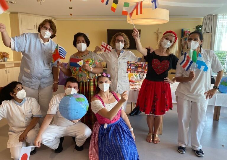 Acht Mitarbeiter*innen der Station Christoph, Seeböckgasse, stehen für ein Gruppenbild zusammen. Einige von ihnen tragen eine Tracht aus ihrem Herkunftsland. Alle schwenken eine kleine Fahne und ein Mitarbeiter hält einen Globus in der Hand.