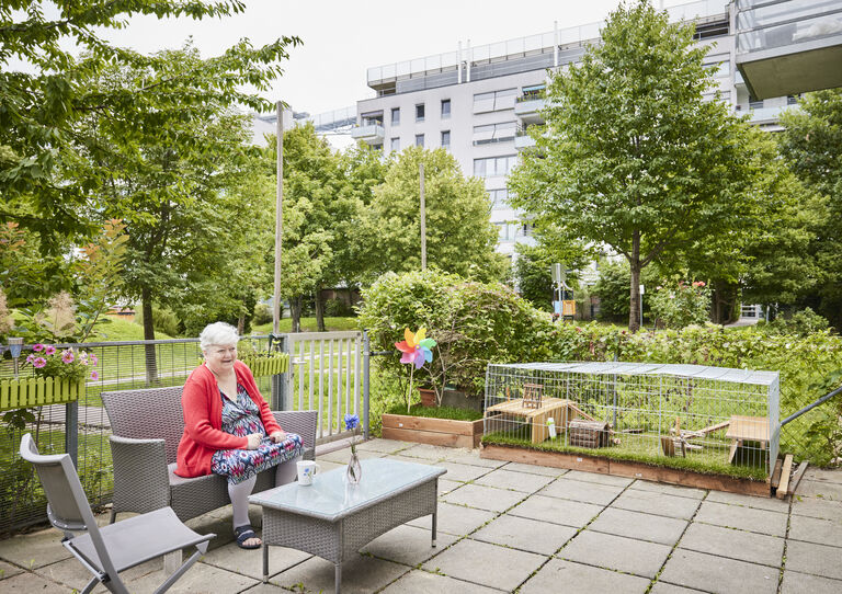 Eine Kundin sitzt im Garten der HABIT WG Thürnlhofstraße an einem Tisch, im Hintergrund ist ein Meerschweinchenkäfig 