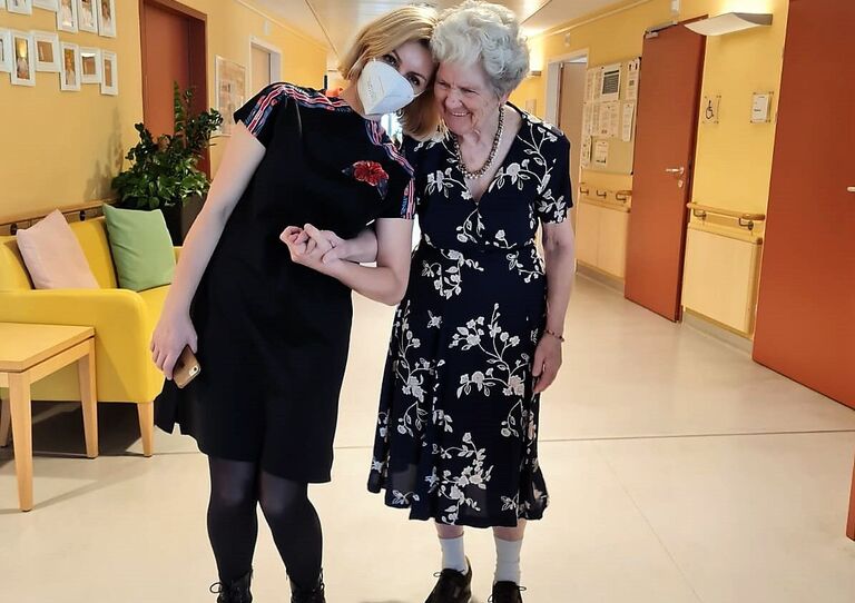 Eine Bewohnerin hält sich bei einer Mitarbeiterin im Stehen ein. Beide haben die Köpfe ganz nahe zueinander und die Mitarbeiterin lächelt.